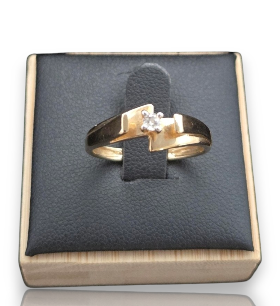 A gold ring with a single clear gemstone displayed on a black textured ring pad inside a light wood box. 