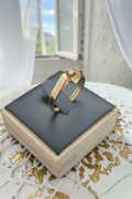 A gold ring with a small clear gemstone displayed on a dark gray cushion atop a light wood box, set on a white lace surface with a window and white curtains in the background. 