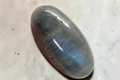 A polished oval labradorite stone with a smooth, rounded cabochon shape, displaying a gray base color with vibrant blue and purple iridescent flashes, resting on a textured light gray surface. 