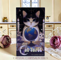 Crystal Chrysalis Haus Cat Tarot card deck box standing upright on a tabletop, featuring a black-and-white cat gazing at a globe with candles, tarot cards, and moon phases in the background. Decorative pink flowers and lace sit around it in a softly lit indoor setting.