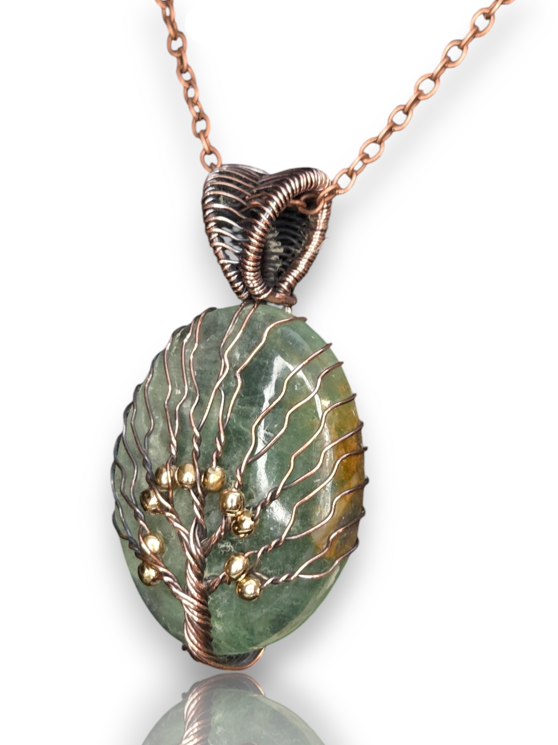 A handmade pendant necklace featuring a green stone cabochon wrapped with copper wire in a tree-of-life pattern, accented with small gold beads, on a white background with a reflection.