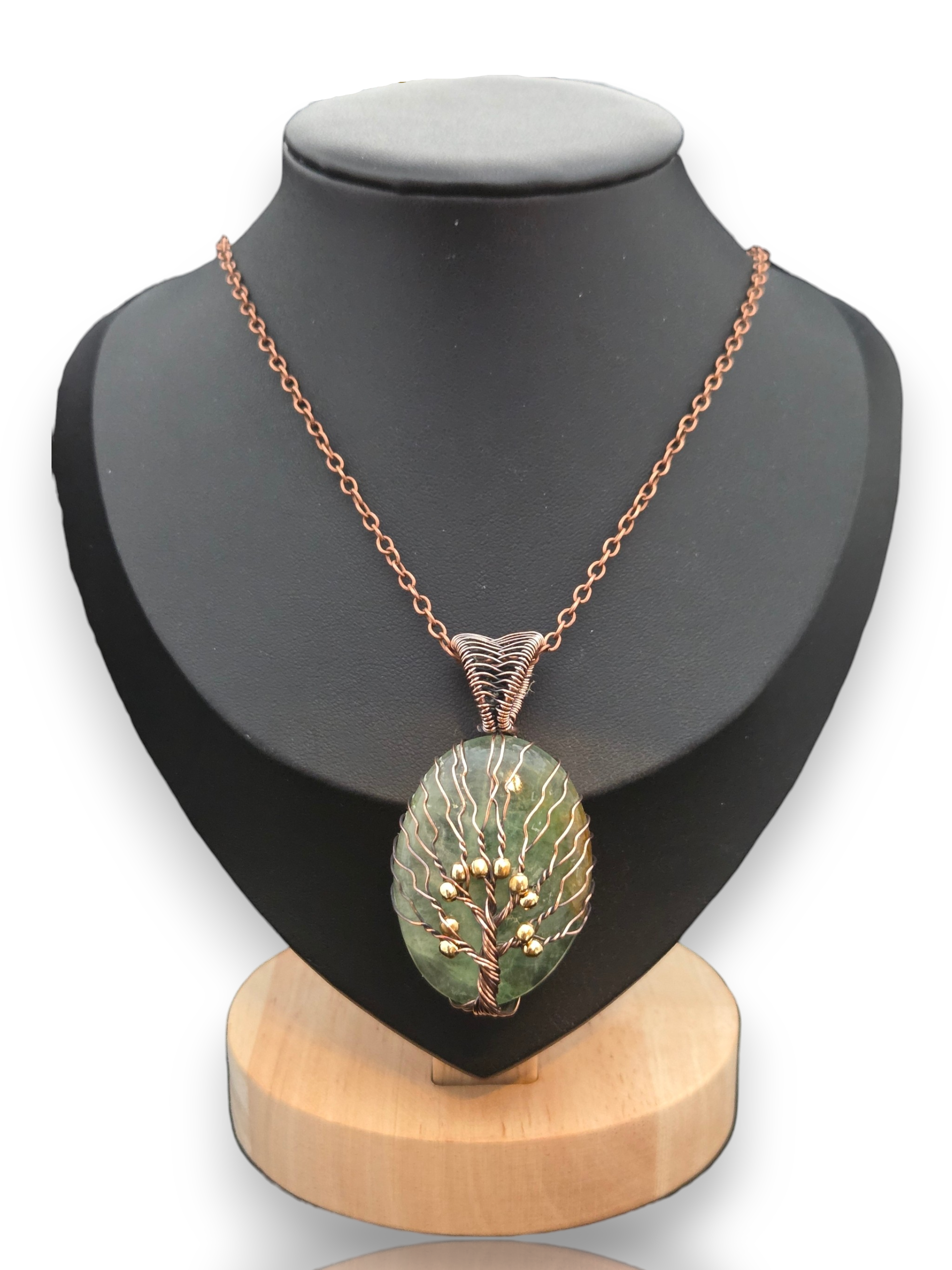 A copper chain necklace with an oval pendant featuring a green and white tree design and small gold berry accents, displayed on a black mannequin bust with a wooden base. 