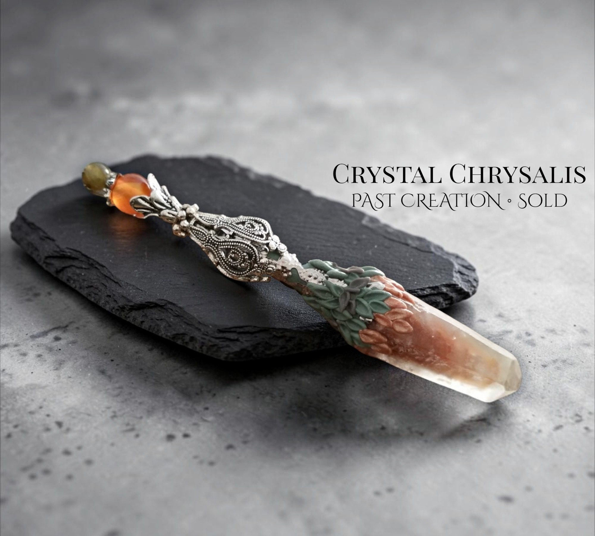 A decorative crystal chrysalis pendant or brooch with an ornate silver-toned filigree top, a small amber bead, and a translucent peach-toned crystal set in green leaf-like detailing, displayed on a dark slate against a gray textured background. Text on the image reads 'CRYSTAL CHRYSALES PAST CREATION - SOLD'.