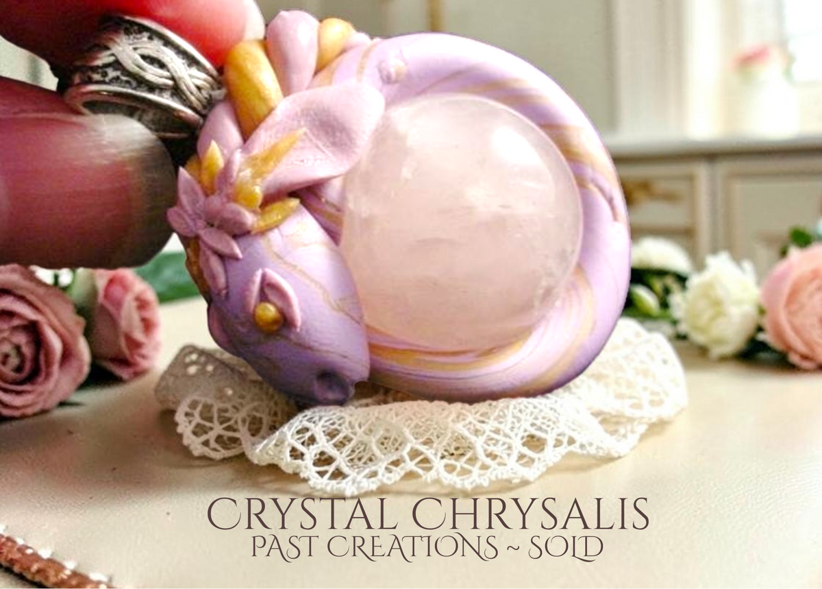 A hand holds a custom dragon handmade crystal chrysalis rose quartz sphere pendant with a lavender and pink swirl body, a pale pink central crystal sphere, and small gold accents, resting on a white lace doily. The background shows a softly blurred indoor setting with pink roses and the text 'CRYSTAL CHRYSALES PAST CREATIONS ~ SOID' at the bottom.