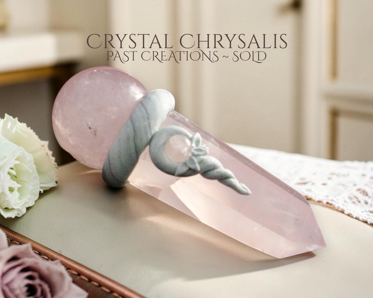 A rose quartz crystal chrysallis point with a carved spiral design wrapped around its center, resting on a beige surface with decorative white and pink roses in the background. Text overlay reads 'CRYSTAL CHRYSLALIS PAST CREATIONS ~ SOLD'.