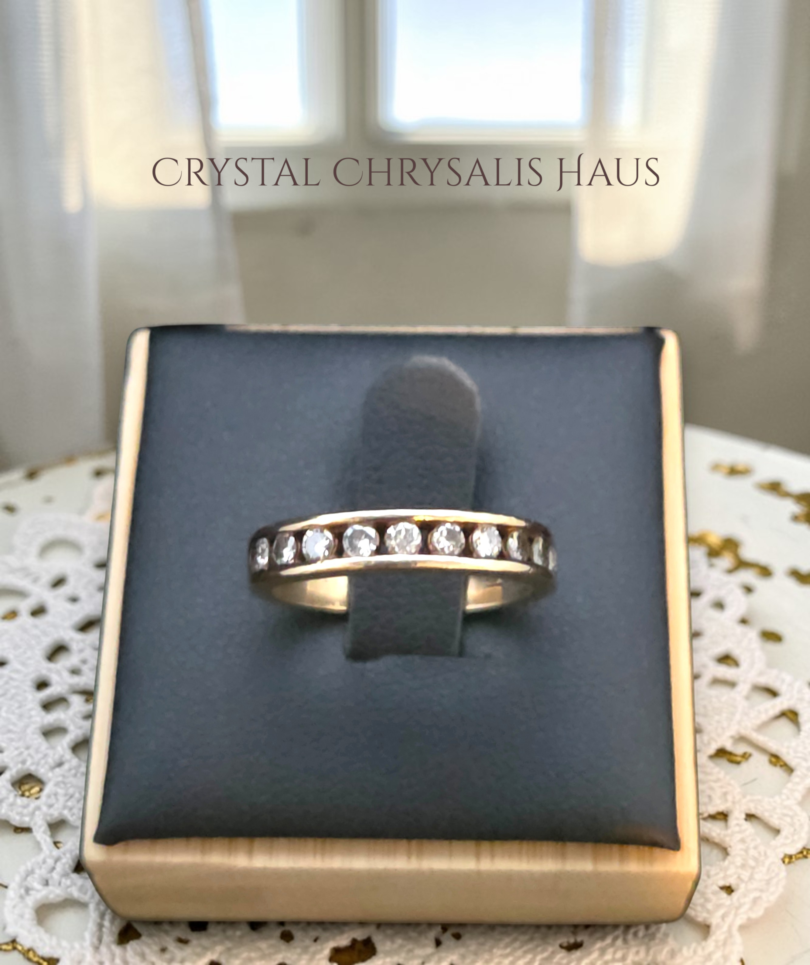 Crystal Chrysalis Haus A silver band ring with a row of diamonds, displayed on a dark gray velvet stand inside a gold-toned jewelry box. The background shows a softly blurred window and sheer curtains, with a white lace doily on the table.