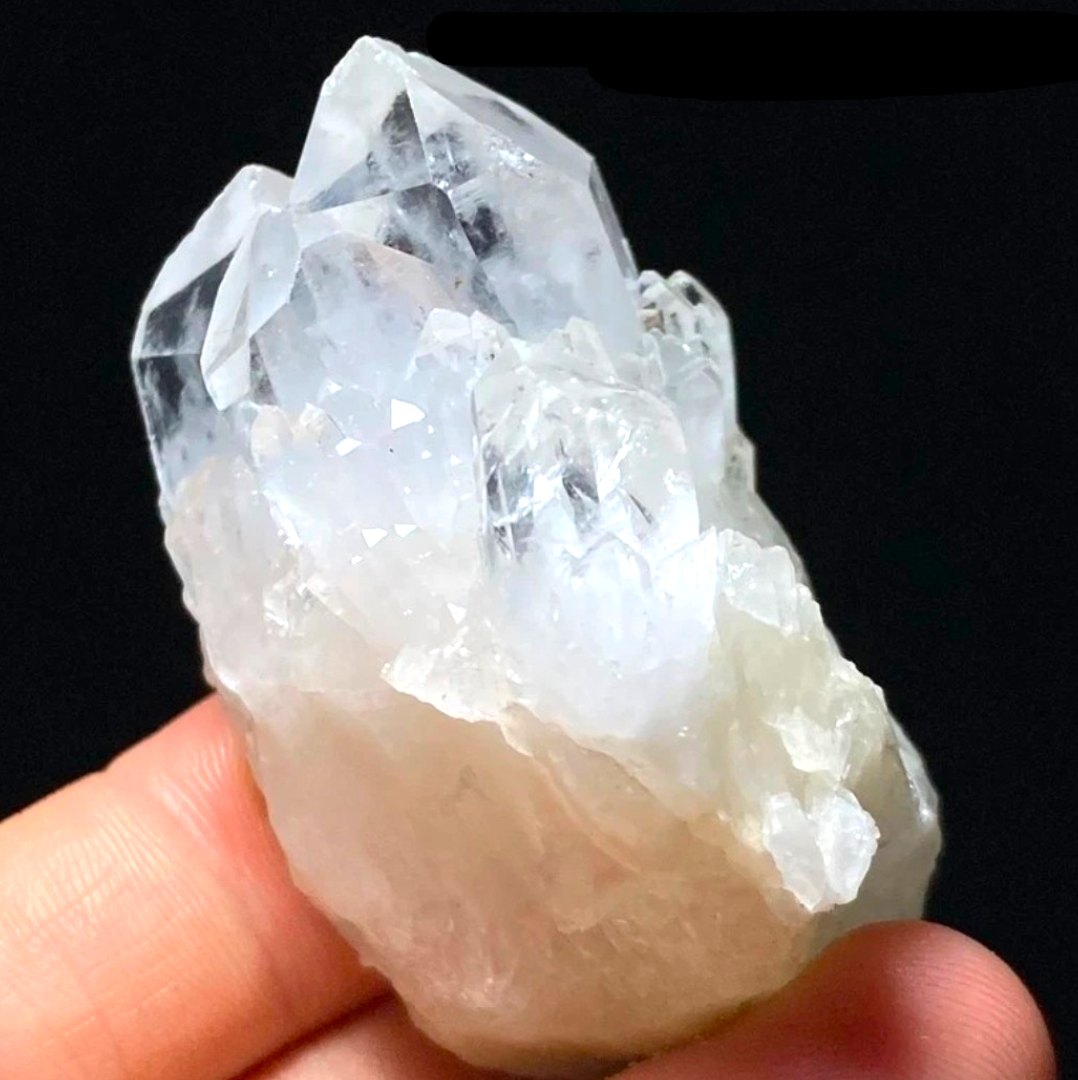 Crystal Chrysalis Haus  A hand holding a raw, rough crystal cluster with multiple clear and semi-translucent points against a solid black background