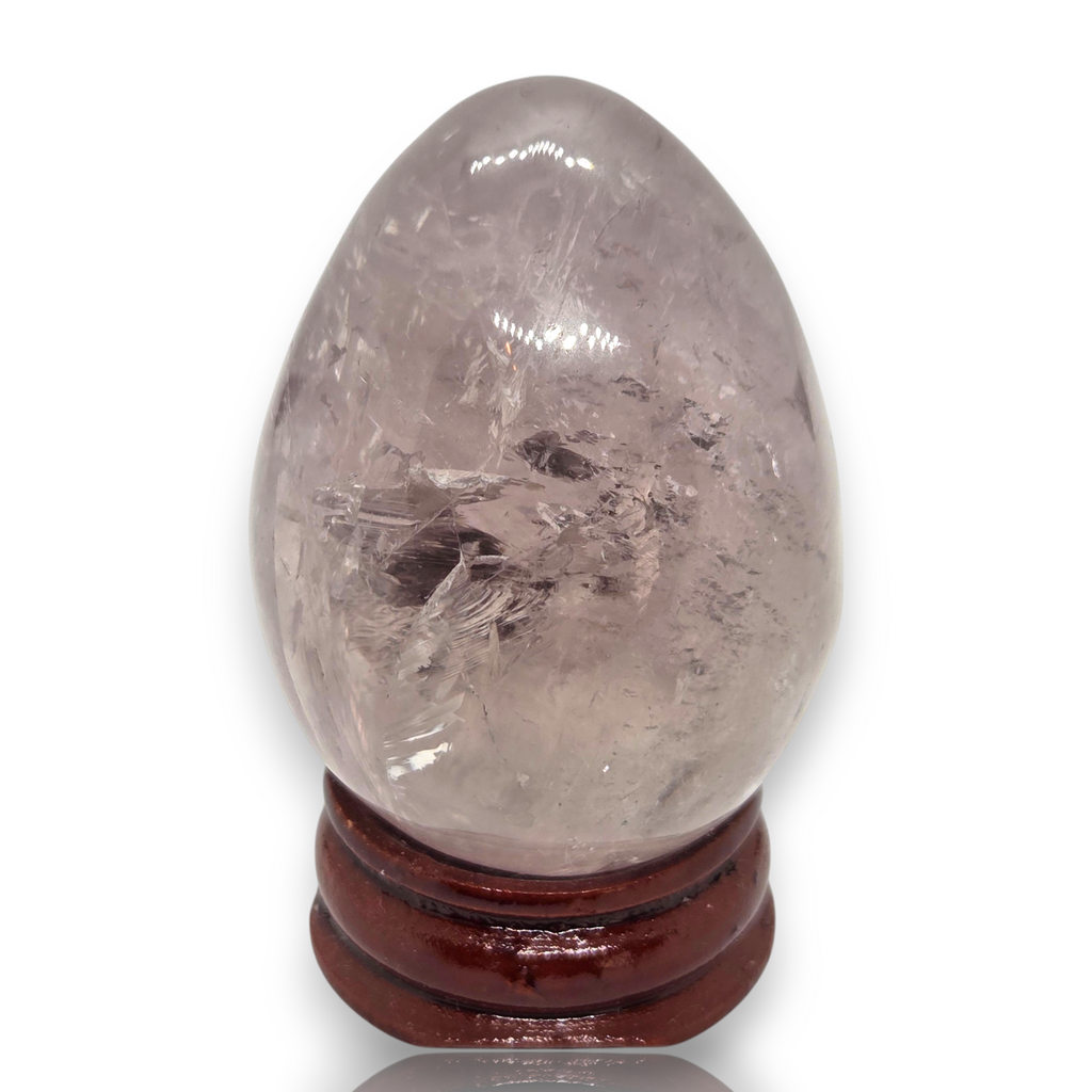 Crystal Chrysalis Haus  A polished light gray quartz crystal egg with visible internal needle-like inclusions, displayed on a dark brown wooden stand against a white background