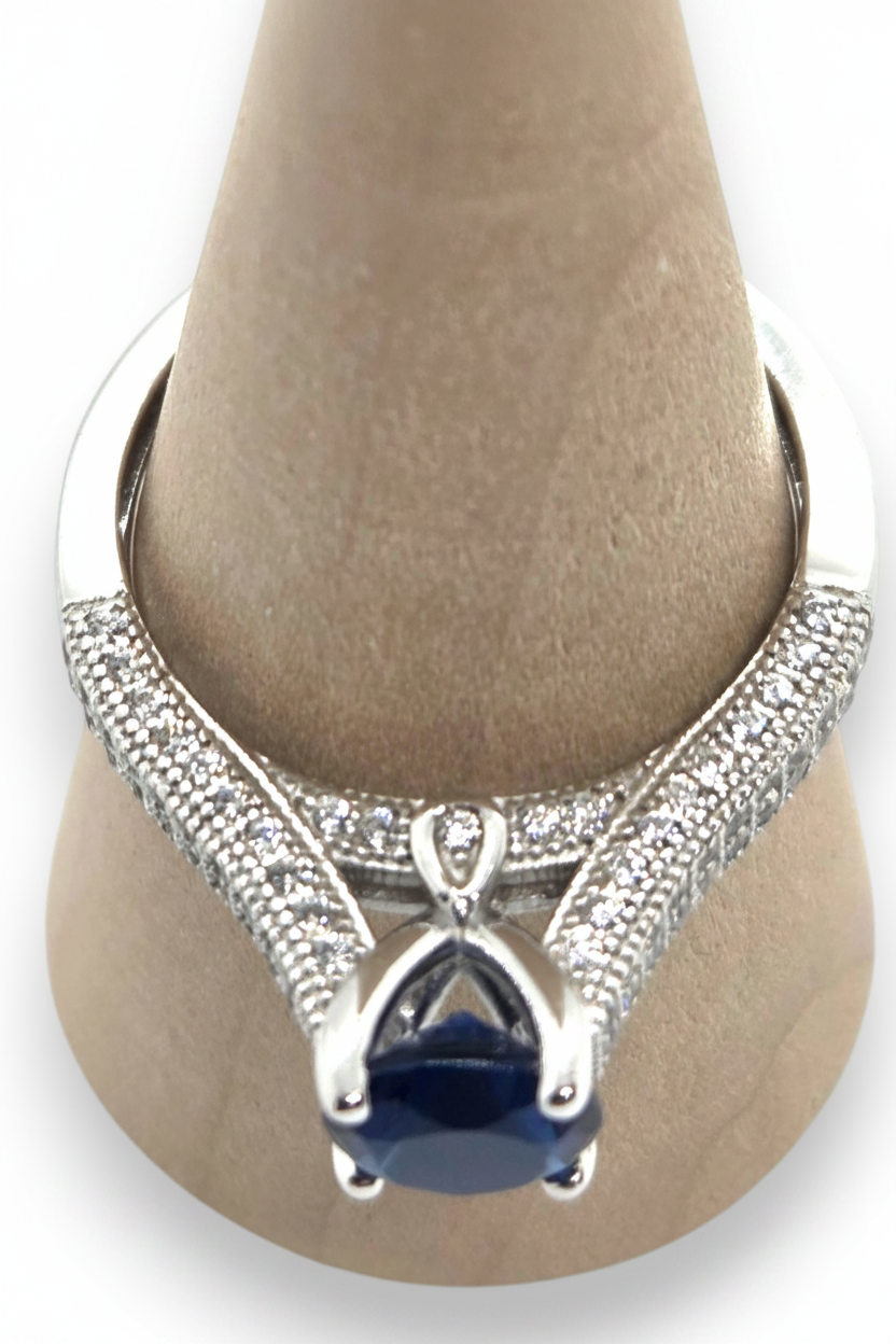 Crystal Chrysalis Haus  A silver ring with a large dark blue pear-cut center stone and pavé-set side stones, displayed on a tan jewelry cone. 