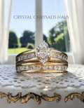 A gold ring set with marquise-cut center stone and rows of clear stones, displayed on a lace doily in front of a window. The text 'Crystal Chrysalis Haus ' is visible at the top.