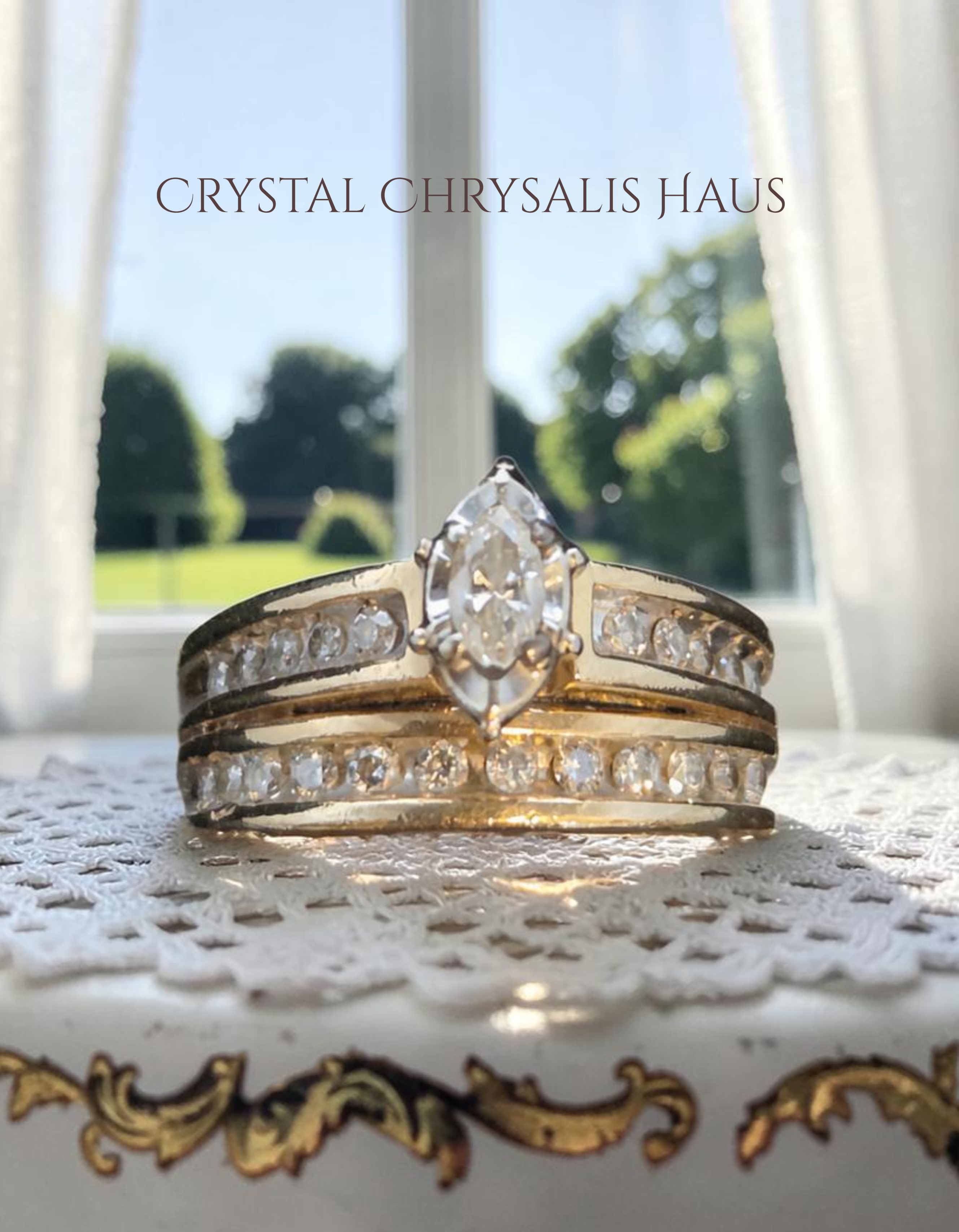 A gold ring set with marquise-cut center stone and rows of clear stones, displayed on a lace doily in front of a window. The text 'Crystal Chrysalis Haus ' is visible at the top.