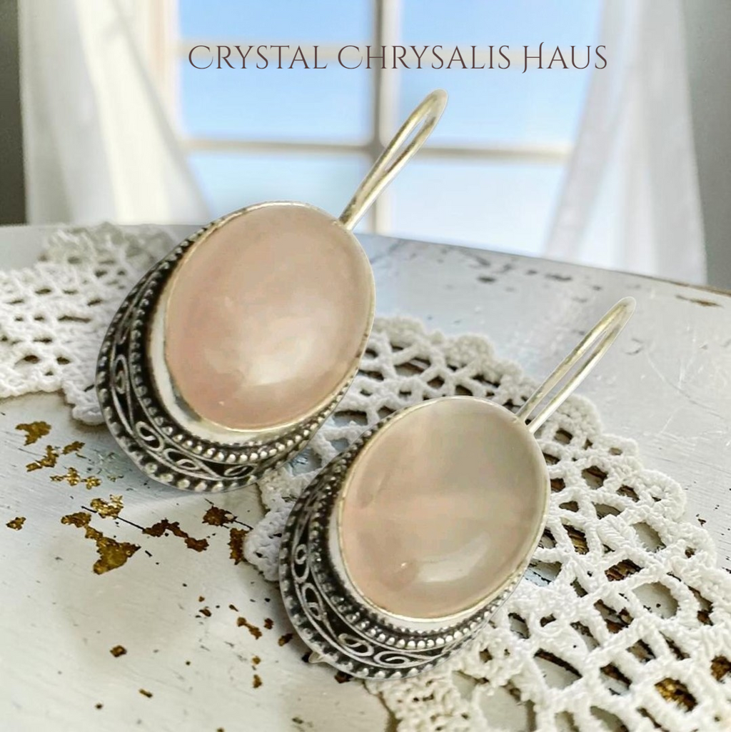 Crystal Chrysalis Haus A pair of dangling earrings with oval pale pink stones set in ornate silver-toned filigree frames, displayed on a white lace doily with a distressed white tabletop and soft window light in the background. The text 'CAT'S ASS BOUTIQUE' appears at the top.