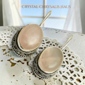 Crystal Chrysalis Haus A pair of dangling earrings with oval pale pink stones set in ornate silver-toned filigree frames, displayed on a white lace doily with a distressed white tabletop and soft window light in the background. The text 'CAT'S ASS BOUTIQUE' appears at the top.