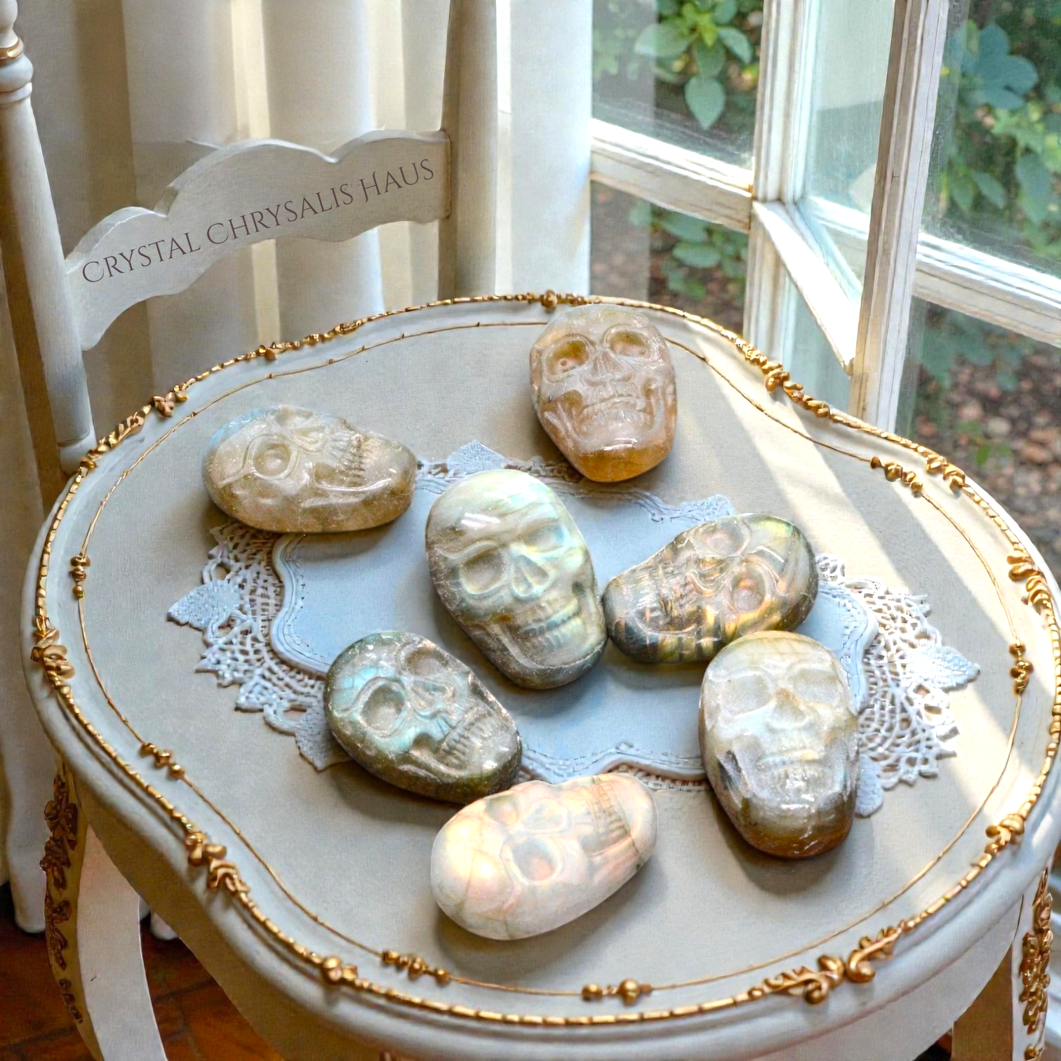 Crystal Chrysalis Haus Seven polished crystal skull stones in various colors and patterns are arranged on a light gray upholstered, gold-trimmed chair in a sunlit room near a window.