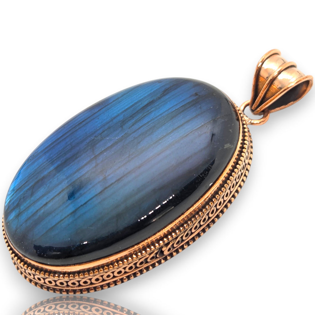 Crystal Chrysalis Haus An oval pendant featuring a vibrant blue labradorite stone with natural striations, set in an ornate rose gold filigree bezel with a matching rose gold bail, shown on a white background with a soft reflection.