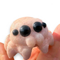 A pink cat-shaped carved object with three glossy black eyes, shown being held in human hands against a white background