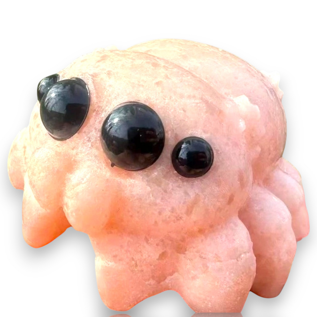 A pink spider-shaped crystal or stone sculpture with three large glossy black bead eyes, shown on a plain white background