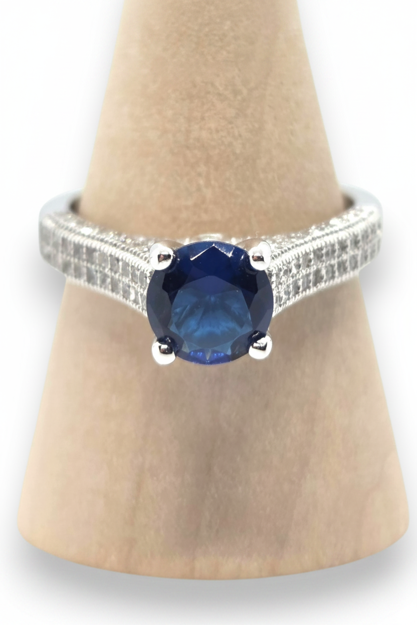 Crystal Chrysalis Haus A silver ring with a round blue sapphire center stone and a pavé-set diamond band, displayed on a tan cone stand. 