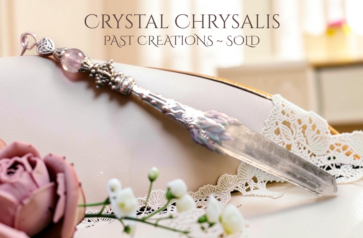 A silver-toned ornate long natural lemurian clear striated crystal point pendant pendulum with a amethyst crystal bead, displayed on a cream surface with lace, a pink rose, and small white flowers. Text at the top reads 'CRYSTAL CHRYSLALIS PAST CREATIONS ~ SOLD'.