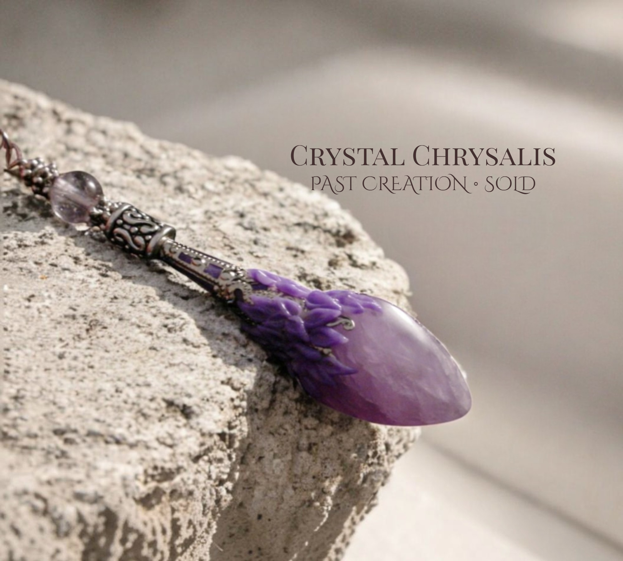 A purple crystal chrysanthemum pendant with silver filigree accents, resting on a textured stone surface. The text 'CRYSTAL CHRYSAALIS PAST CREATION - SOLD' is overlaid on the upper right.