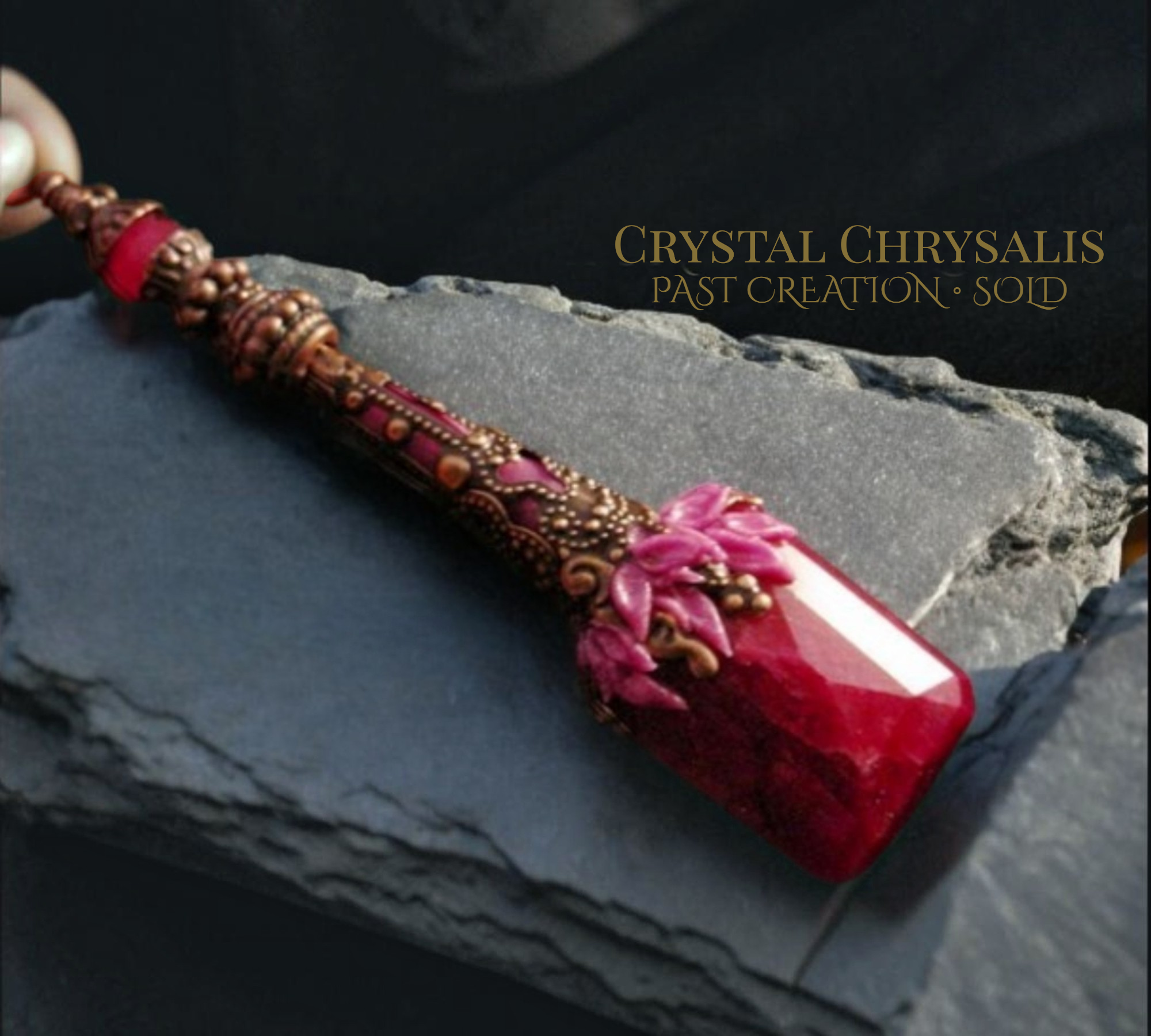A handmade crystal chrysalis pendant with a faceted deep red gemstone, ornate copper-toned metalwork, and pink flower details, resting on a dark slate surface with text overlay reading 'CRYSTAL CHRYSalIS PAST CREATION • SOLD'.