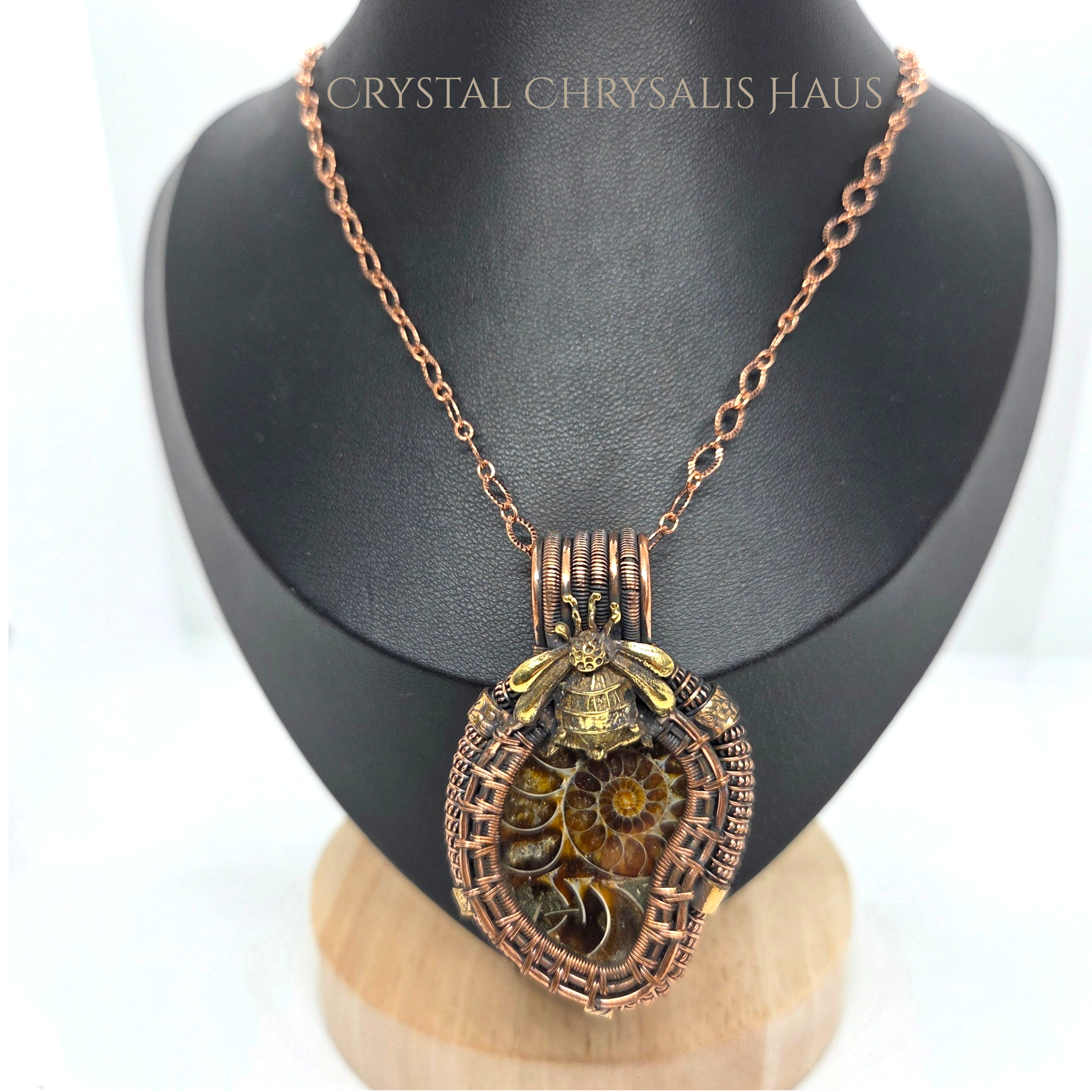 A copper-toned necklace with a detailed mechanical pendant featuring a bee and spiral gears, displayed on a black jewelry bust.