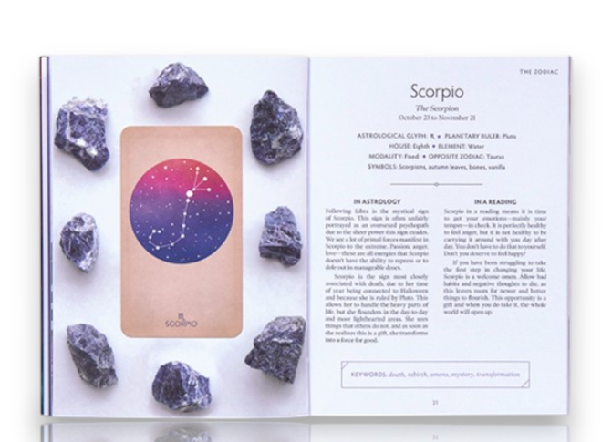 An open astrology-themed booklet or card deck spread and 'Scorpio,' surrounded by several dark purple crystals on a white surface.