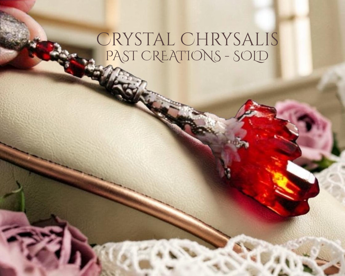 A hand holds a crystal chrysalis pendant with a large rare  red aura crystal cluster and ornate silver-toned metalwork, resting against a cream-colored leather surface with pink roses and lace in the background. Text at the top reads 'CRYSTAL CHRYSalis PAST CREATIONS - SOLD'.