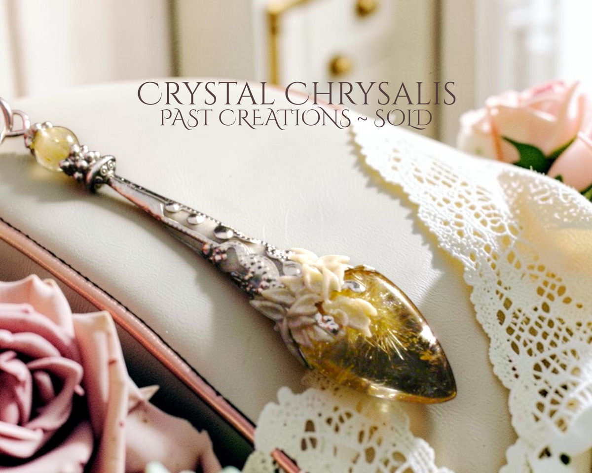 A decorative spoon-like crystal chrysalis pendant with an ornate silver-toned handle and a gold threads rutile crystal pendant, displayed on a cream surface with lace and pink roses. Text overlay reads 'CRYSTAL CHRYRALIS PAST CREATIONS ~ SOLD'.