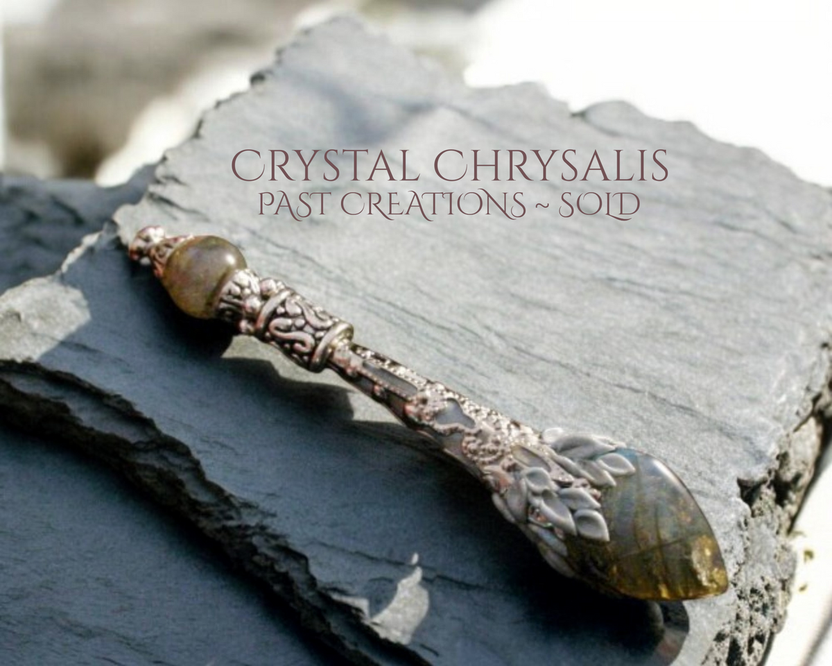 An ornate silver-toned chrysanthemum-shaped crystal pendant with a greenish-brown top bead and a faceted green crystal end, resting on a dark slate rock. Text overlay reads 'CRYSTAL CHRYSalis PAST CREATIONS ~ SOLD'.