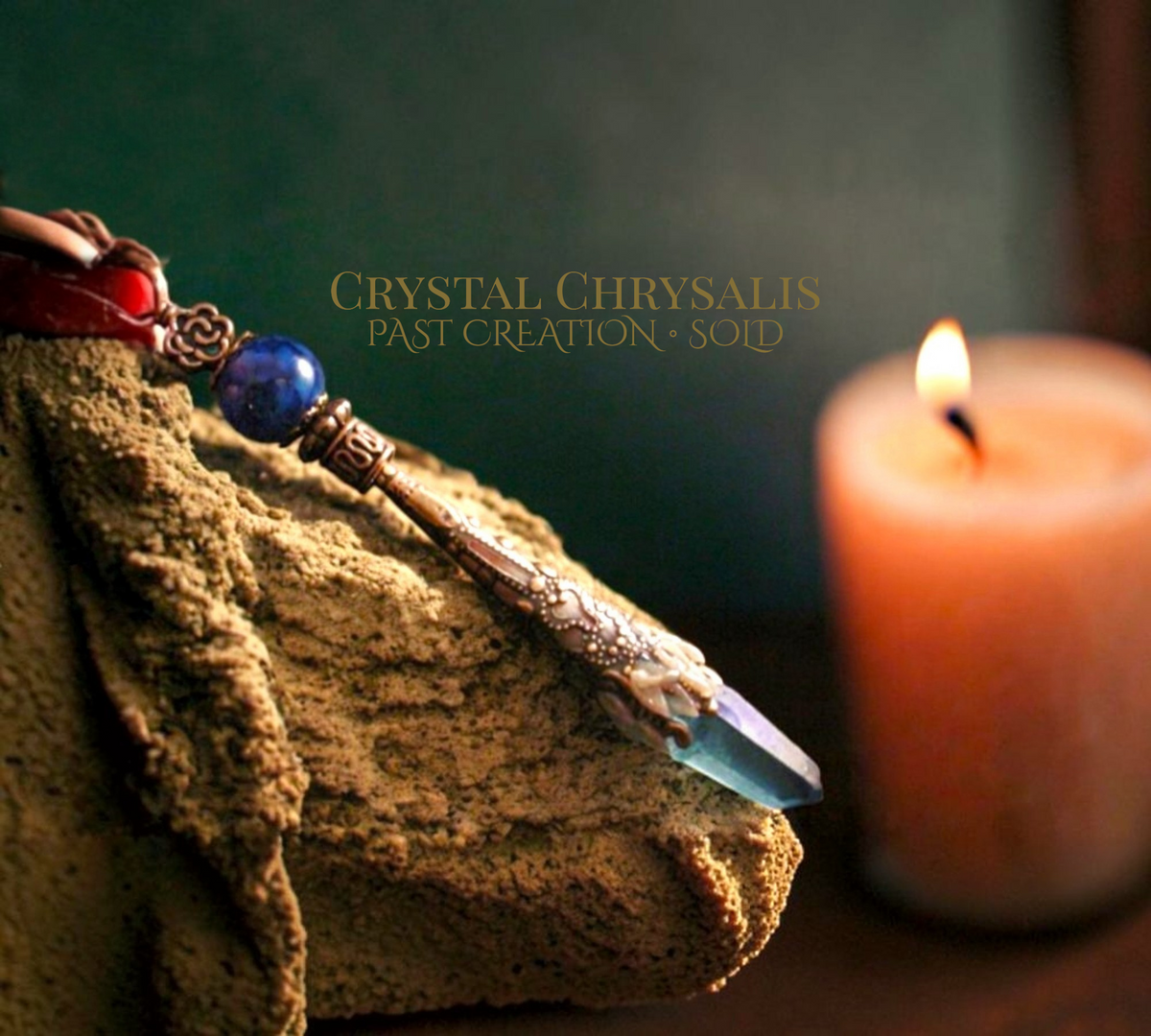 A close-up of a decorative pendant or charm with a faceted blue crystal, ornate metal detailing, and a blue bead, resting on a textured tan cloth beside a lit candle. The text 'CRYSTAL CHRYsalis PAST CREATION - SOLD' appears over the center.