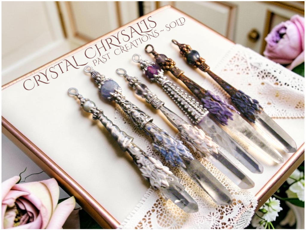 Six Custome handmade quartz crystal point pendants & pendulums with ornate silver and copper filigree fittings are arranged on a cream-colored presentation box with the text 'CRYSTAL CHRYSALIS  PAST CREATIONS - SOLD'. The box is decorated with a white lace runner and pink roses are visible in the background and foreground.