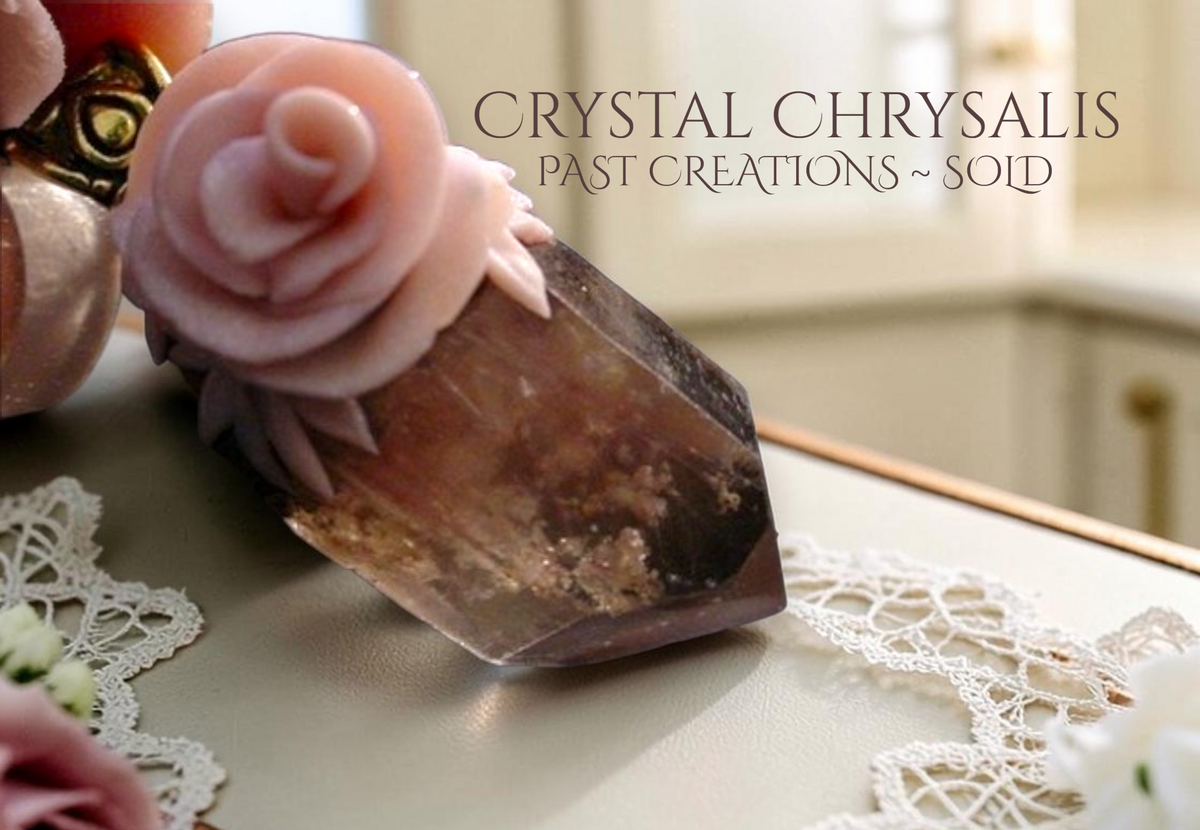 A polished crystal chrysalis with a pink rose accented smoky inner scene quartz crystal point pendant, displayed on a beige surface with white lace doilies and soft-focus background decor. Text overlay reads 'CRYSTAL CHRYSA LIS PAST CREATIONS ~ SOLD'.