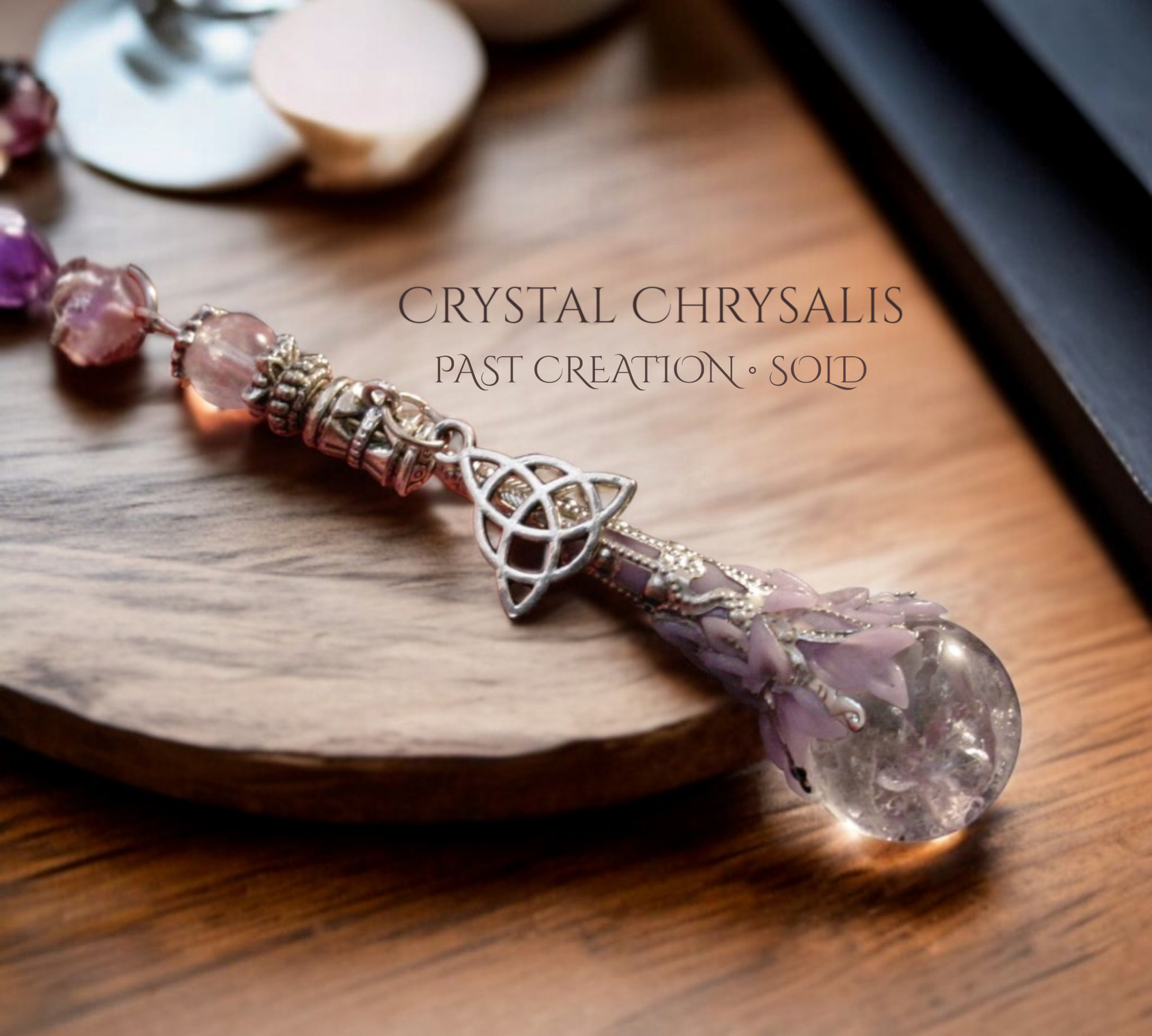 A crystal chrysalis pendant with a large clear teardrop-shaped crystal, purple dried flower inclusions, and a silver Celtic knot charm, resting on a wooden surface with text overlay 'CRYSTAL CHRYSAIS PAST CREATION • SOLD'.