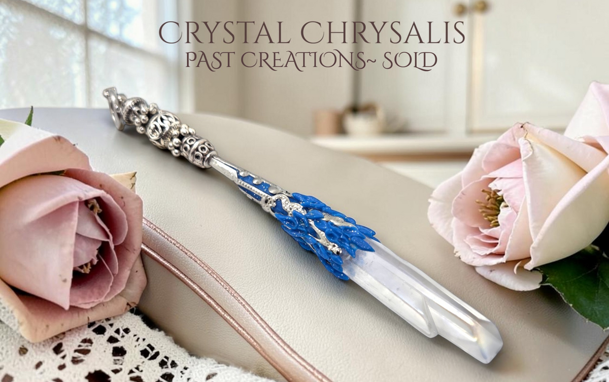 A decorative crystal chrysalis pendant or keepsake lies on a beige leather surface, styled with pale pink roses and lace. The object has an ornate silver-toned filigree cap, a bright blue textured body, and a angel aura white crystal .