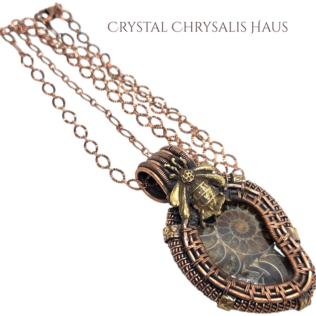 A copper and brass wire-wrapped pendant necklace featuring a fossilized ammonite shell inside a Greek key border, with a small gold beetle charm, shown on a white background with the text 'CRYSTAL CHRYsalis HAUS' at the top.