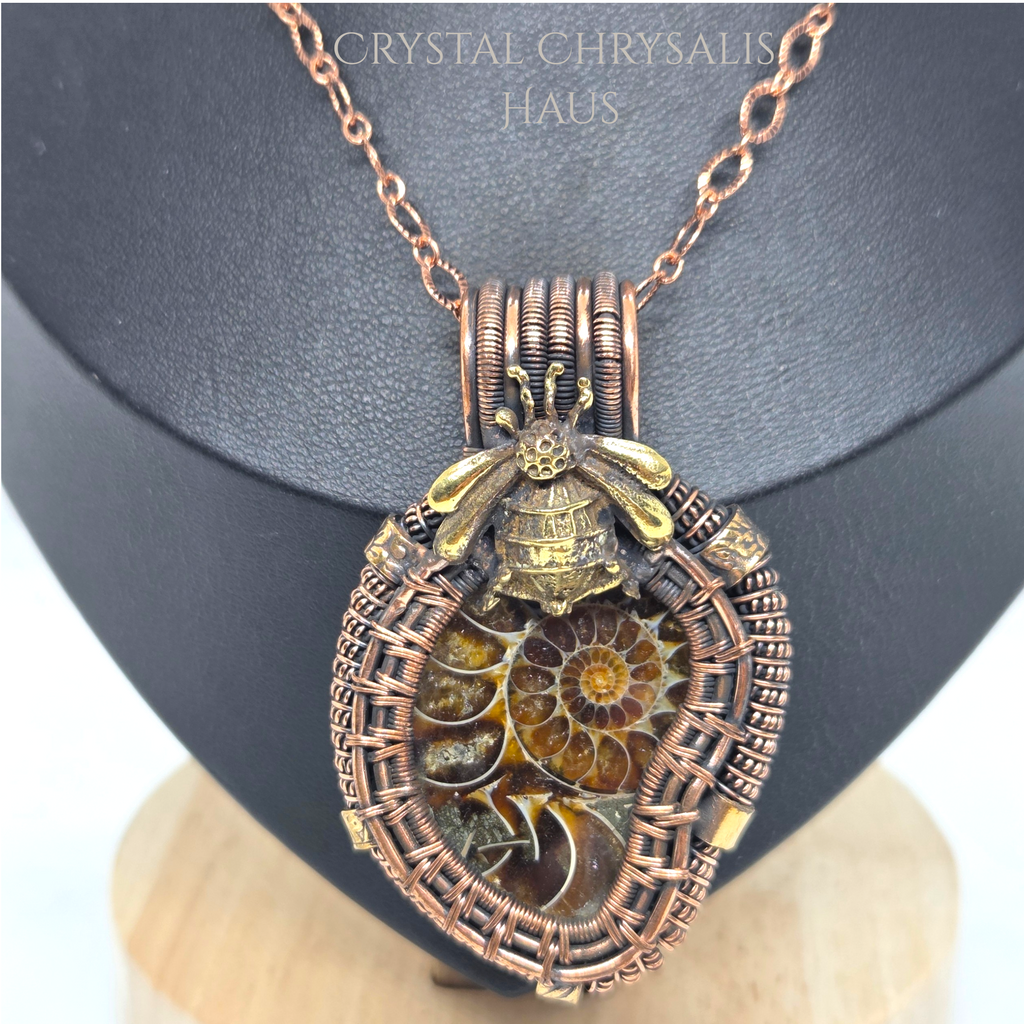 A copper-wrapped pendant necklace featuring a fossilized ammonite shell and a small gold-toned bee charm, displayed on a black jewelry bust.
