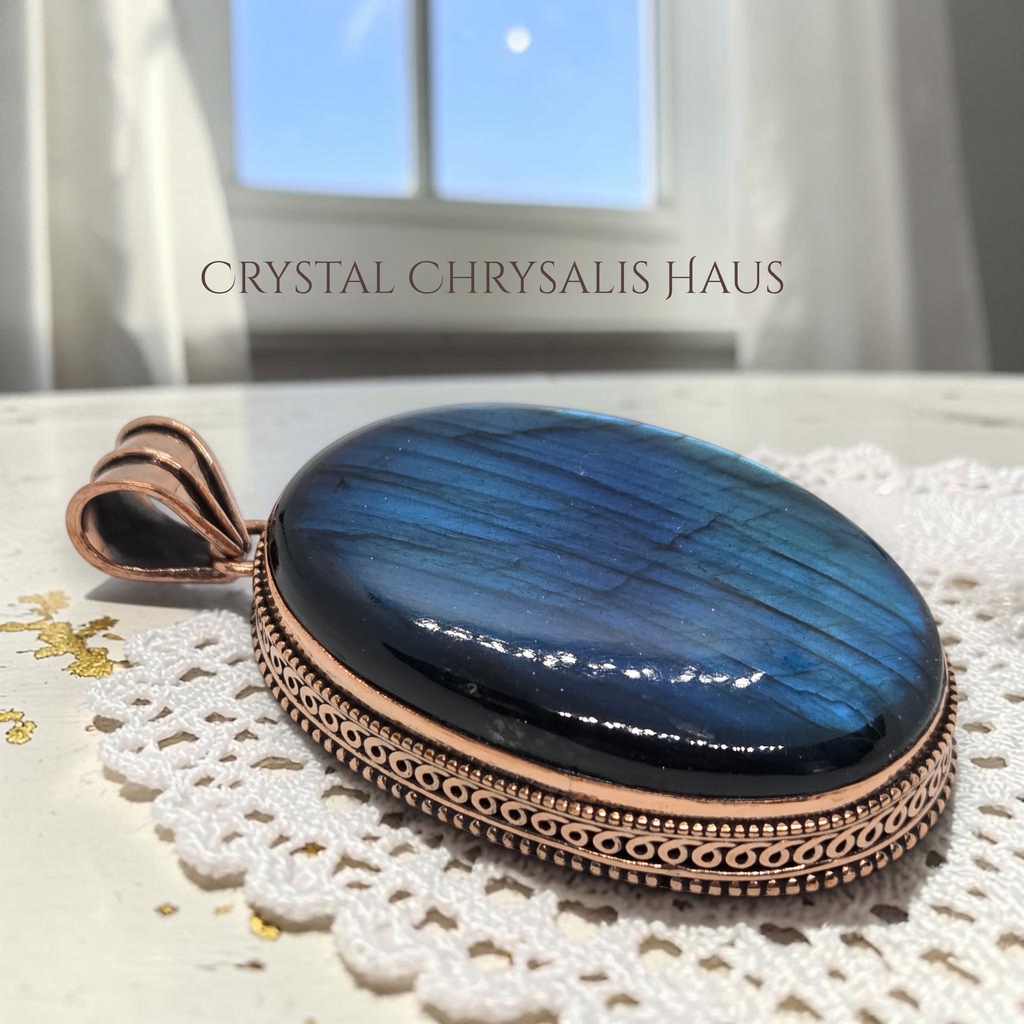 Crystal Chrysalis Haus A polished oval pendant with a deep blue iridescent stone and a rose gold-toned metal border, displayed on a white lace doily with a window in the background