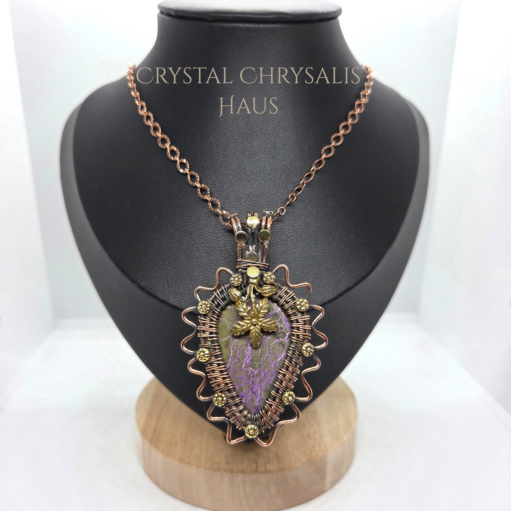 A crystal chrysalis pendant necklace displayed on a black jewelry bust. The pendant features a purple and green chrysalis insect encased in a copper wire frame with small gold beads, suspended from a rose gold chain.