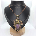 A crystal chrysalis pendant necklace displayed on a black jewelry bust. The pendant features a purple and green chrysalis insect encased in a copper wire frame with small gold beads, suspended from a rose gold chain.
