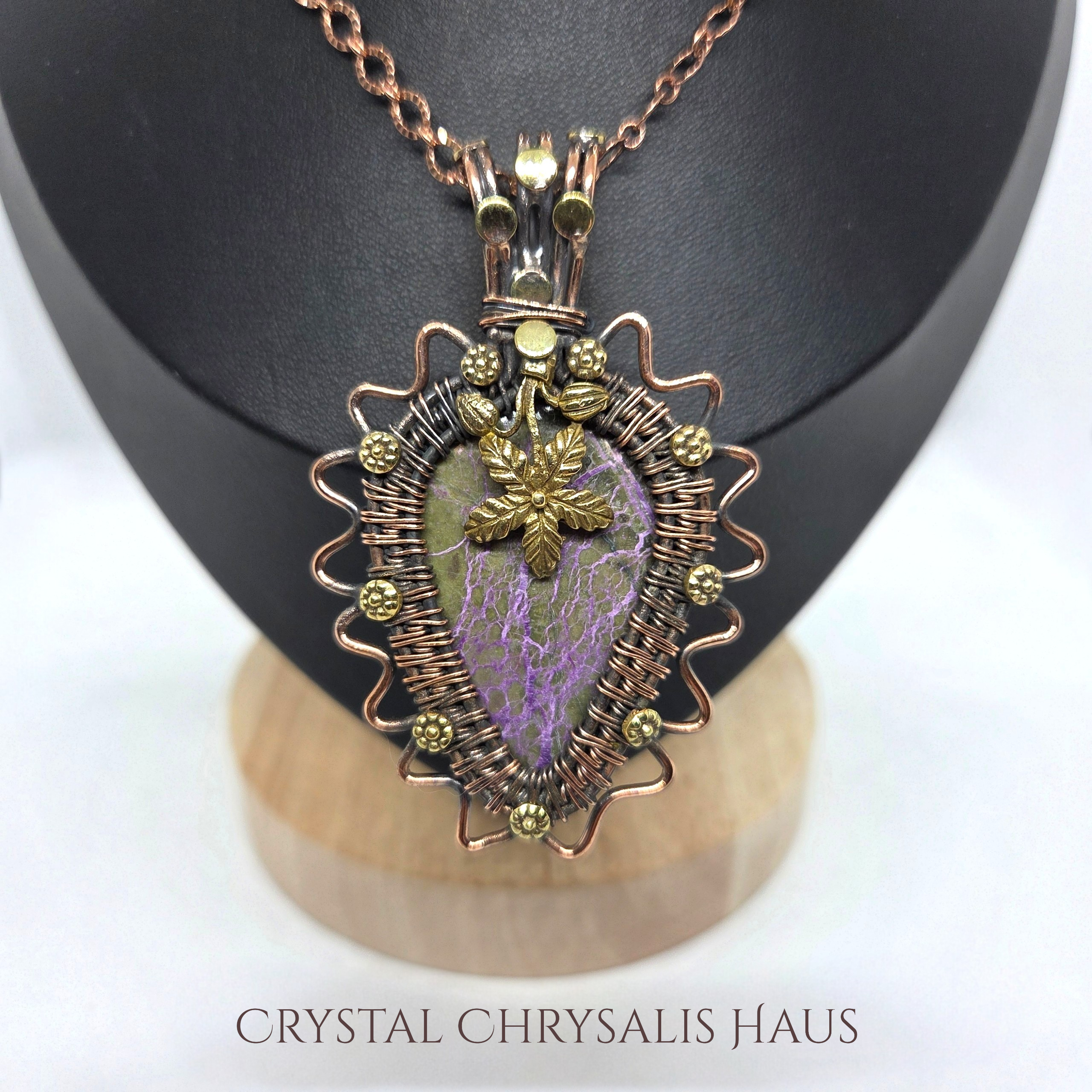 A handmade wire-wrapped pendant necklace featuring a purple and green stone with a gold leaf charm, displayed on a black jewelry bust with a wooden base. The brand name 'CRYSTAL CHRYSLIS HAUS' is printed at the bottom.