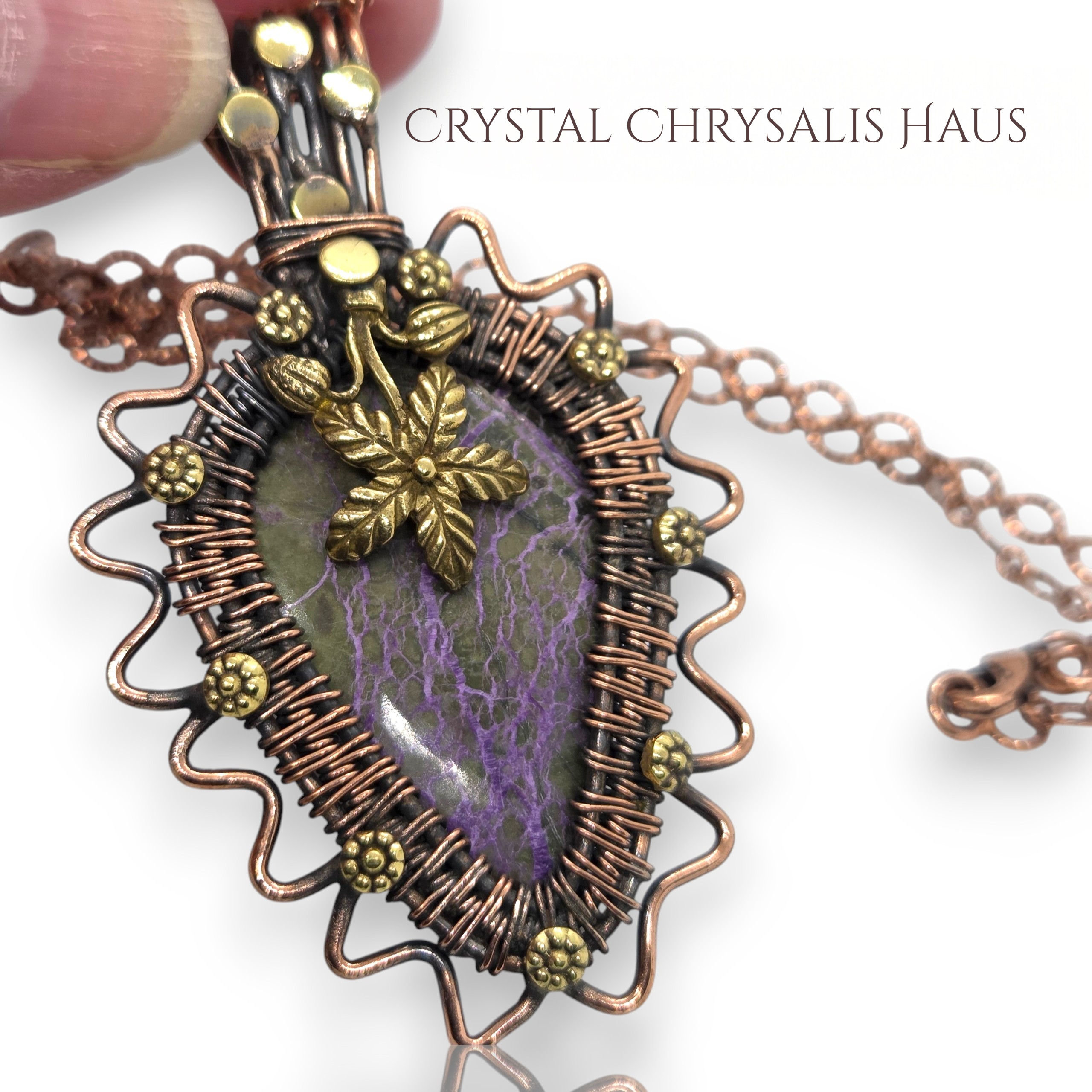 A close-up of a handmade teardrop-shaped pendant necklace with a purple and green stone, wrapped in copper-toned wire with gold-toned accents, held by a hand against a white background 