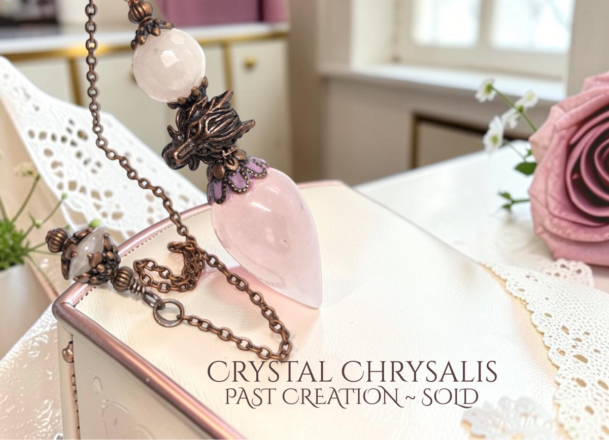 A close-up of a hand holding a bronze chain necklace with a large pale pink crystal pendant and smaller white crystal beads, resting over a white lace-covered surface with a pink rose in the background. Text at the bottom reads 'CRYSTAL CHRYSALIS PAST CREATION ~ SOLD'.