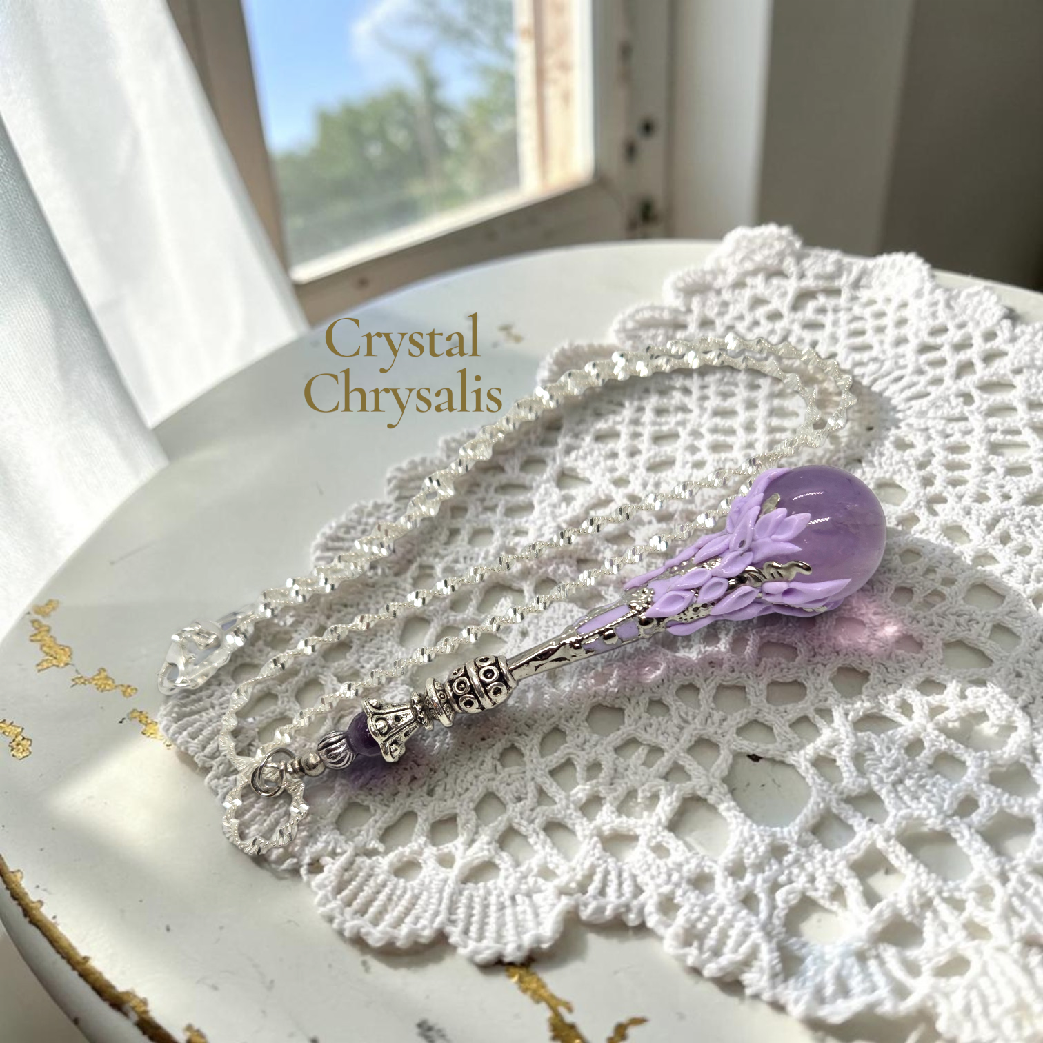 A purple crystal chrysanthis pendant with an ornate silver-toned bail, displayed on a white lace doily next to a strand of clear crystals. The text 'Crystal Chrysalis' is overlaid on the image.