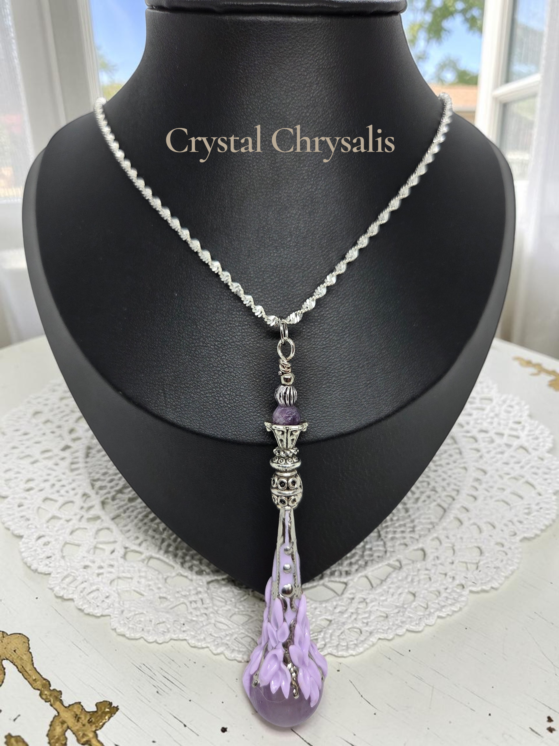 A silver-toned beaded chain necklace with a teardrop-shaped lavender crystal pendant, displayed on a black jewelry bust. The necklace is shown against a background with a white lace doily and a window.
