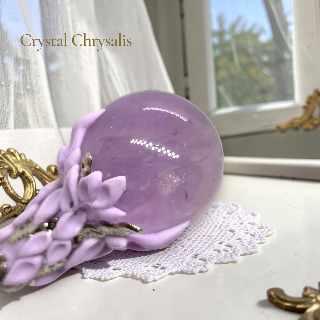 A lavender purple amethyst crystal ball ornament displayed on a decorative lavender floral stand with silver accents, set on a white lace doily in front of a window.